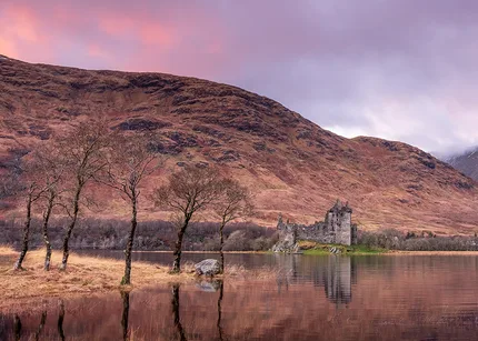 The sky is pink and purple as the sun sets over Kilchurn Castle. The mountain is a backdrop whilst trees on an island reflect into the water.