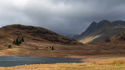 A landscape scene of hill and mountains with lake in front with a golden glow and moody sky.