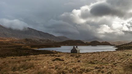 A fishing bothy stands on the edge of Loch Assynt.  The landscape is bleak with thunderous clouds looming over.