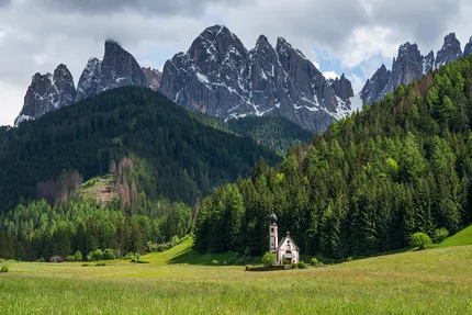 A Baroque church stands alone in a meadow with towering peaks of the Dolomites behind.