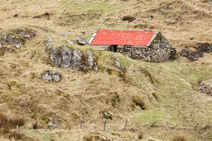 A crofter's cottage with red tin roof stands against thick stone walls against ruffed moorland.