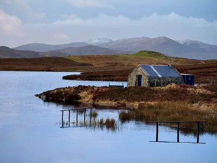 A small fishing bothy sits besides the water edge  surround by rugged wilderness of the Scottish Highlands
