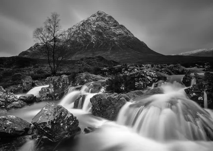 In black and white a waterfall cascades over ancient rocks.  Beyond is mountains of Glen Etive.