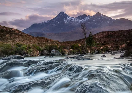 Mountains in Glencoe with pink and purple clouds.  Slow shutters speed water cascades over rocks in the foreground