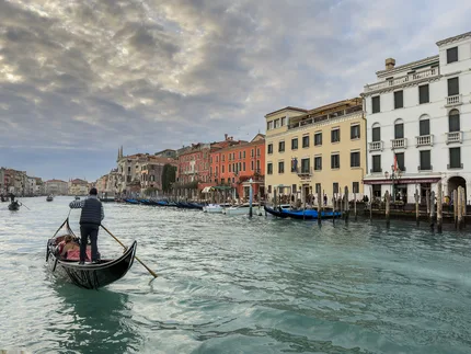 A lone gondola gliding away towards the heart of Venice.  Venetian buildings recede into the distance.  There's a brooding moody sky adding drama to the scene.