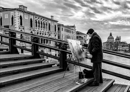 An elderly artist stands on Accademia Bridge, Venice,  painting the landscape.   Captured in black and white