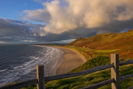 As the light sweeps across Rhossili Bay, the landscape is bathed in a warm golden glow. Waves roll in from the Atlantic Ocean