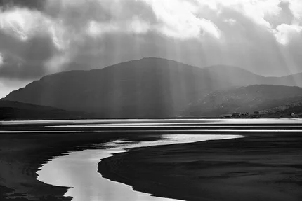 Receding tide leaving ribbon like stream backed by mountains. Sunbeams break through the clouds.