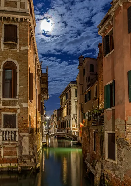 A Venice canal lies beneath the soft glow of the moon.  Ancient buildings rise on either side.